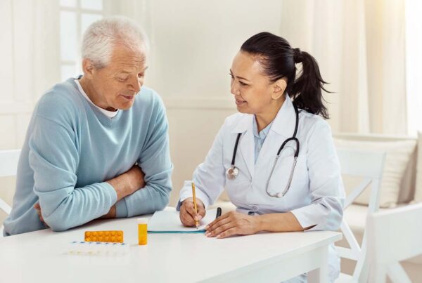 Medicare Part D - Smiling and Encouraging Doctor Takes Notes as She Discusses Medication Precautions and Side Effects with a Senior Male Patient in His Home at the Kitchen Table