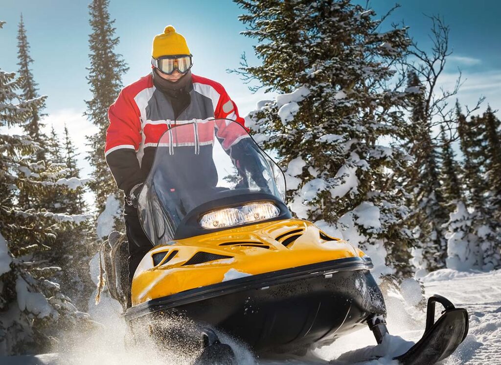 Snowmobile Insurance - Close-up of a Man in Winter Attire and Goggles Riding a Snowmobile Through Snow and Trees on a Winter Day