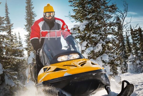 Snowmobile Insurance - Close-up of a Man in Winter Attire and Goggles Riding a Snowmobile Through Snow and Trees on a Winter Day