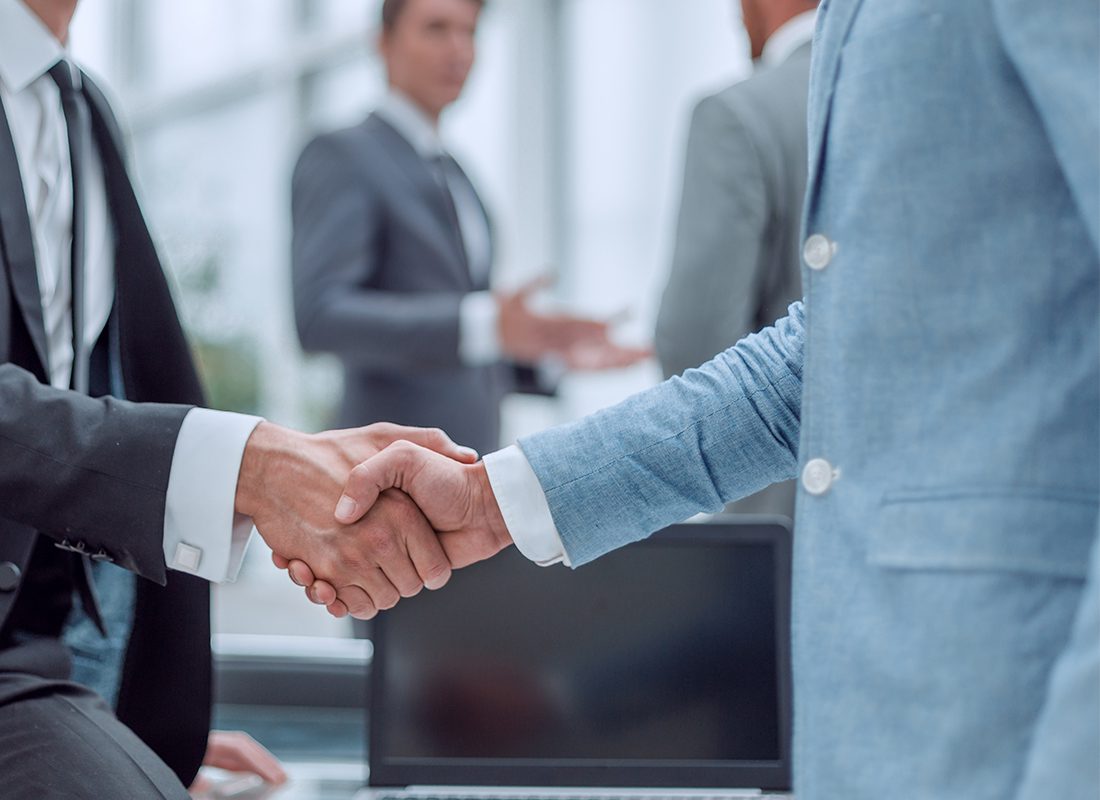 Bob Korvas Agency - Close Up of Two Professionals in Suits Shaking Hands in an Office in Front of a Computer with Other Professionals Discussing in the Background